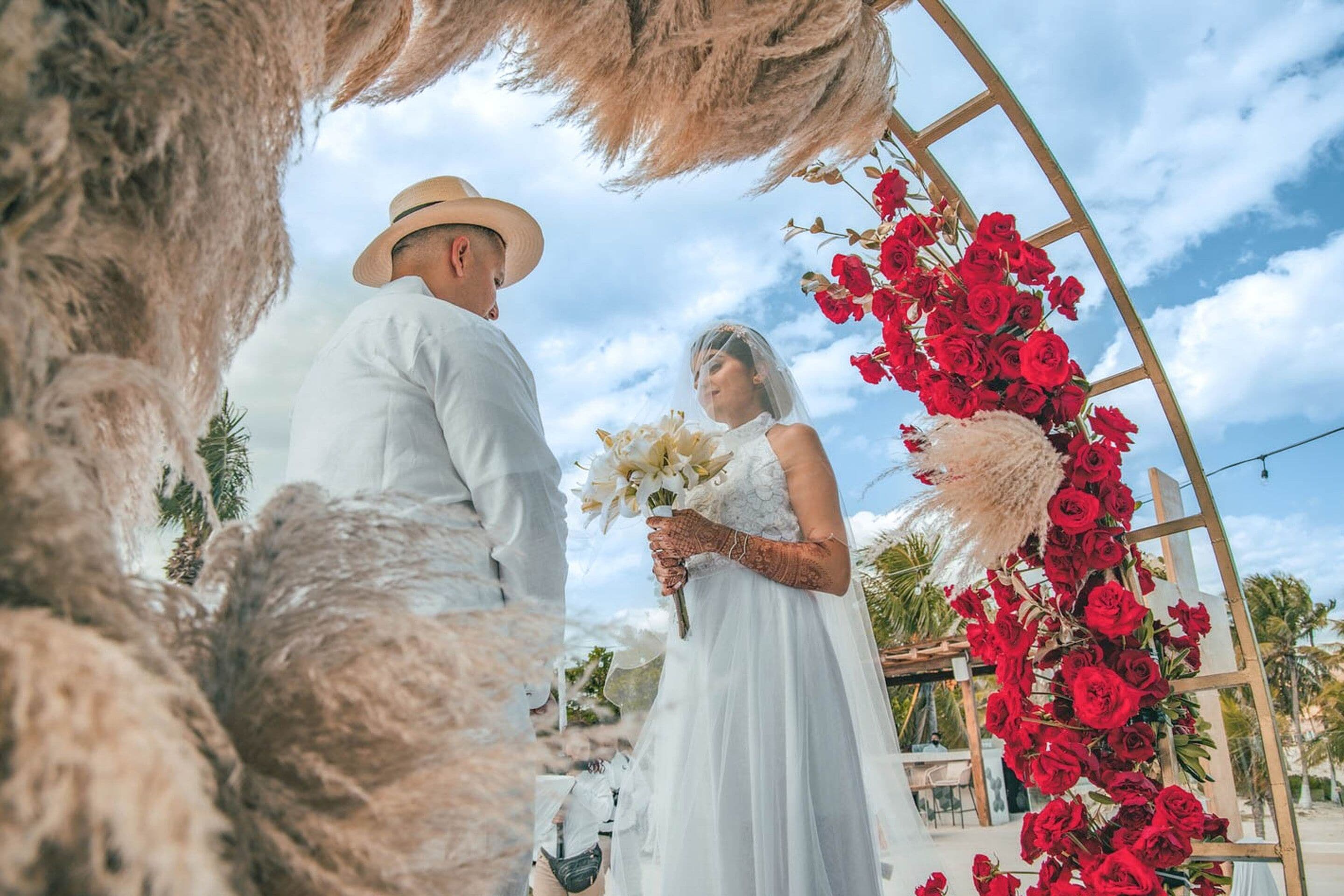 A wedding ceremony with a wooden arch adorned with red roses and pampas grass.