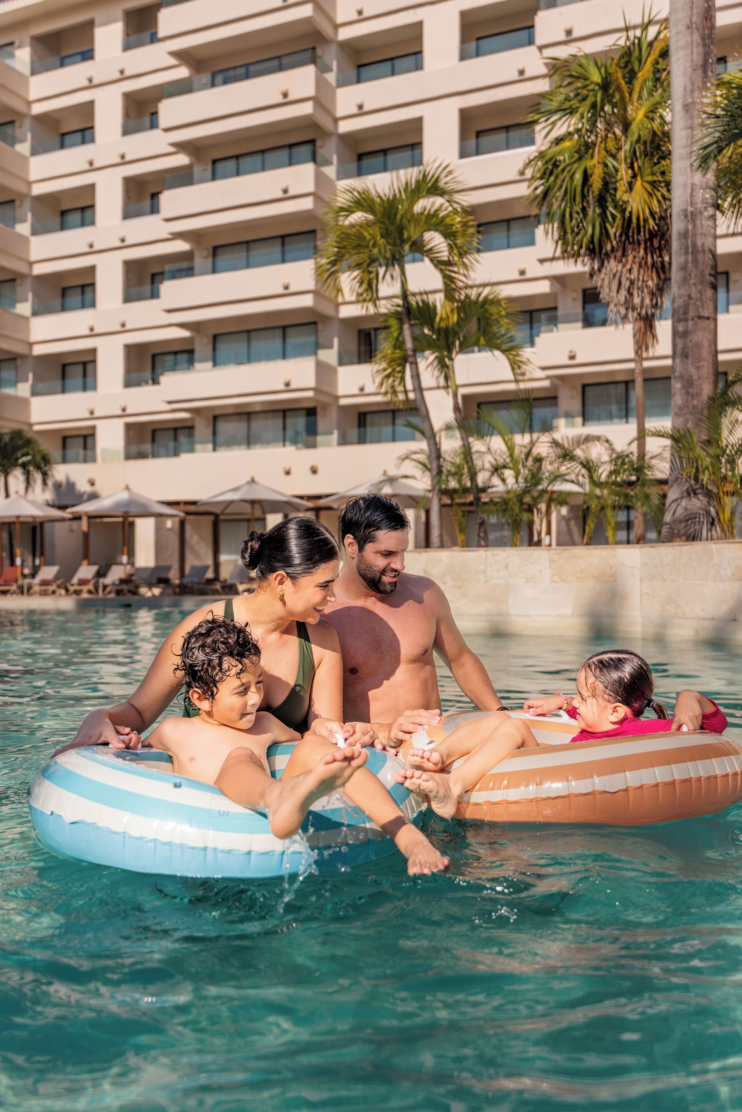 En swimmingpool med oppustelige ringe, palmer og en hotelbygning i baggrunden.