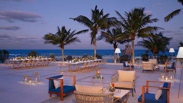 An outdoor dining area with tables and chairs, palm trees, and a view of the ocean.