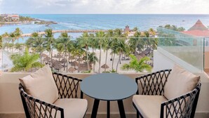 Un balcon avec deux fauteuils en osier et une petite table, donnant sur une plage bordée de palmiers et offrant une vue sur l’océan.