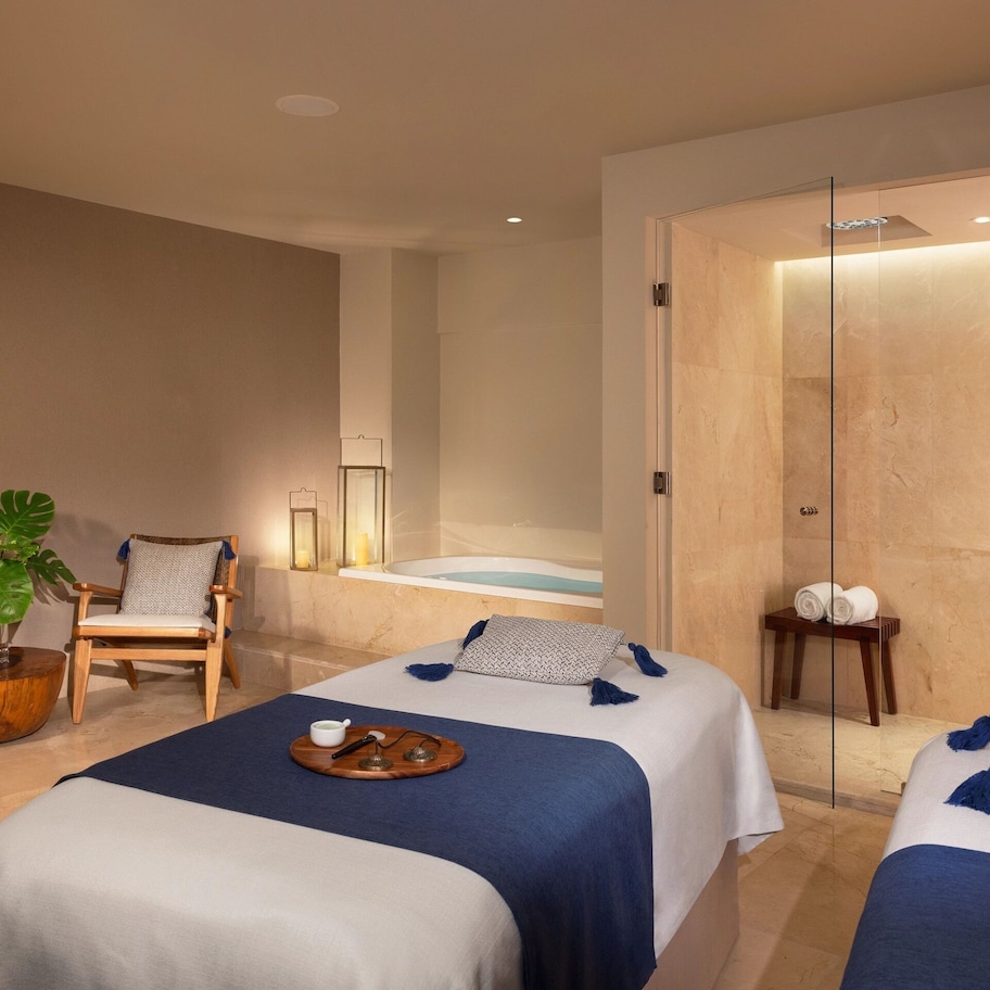 A hotel room with two beds, a sitting area with chairs, a plant, a small table with a tray, and a glass-enclosed shower area.