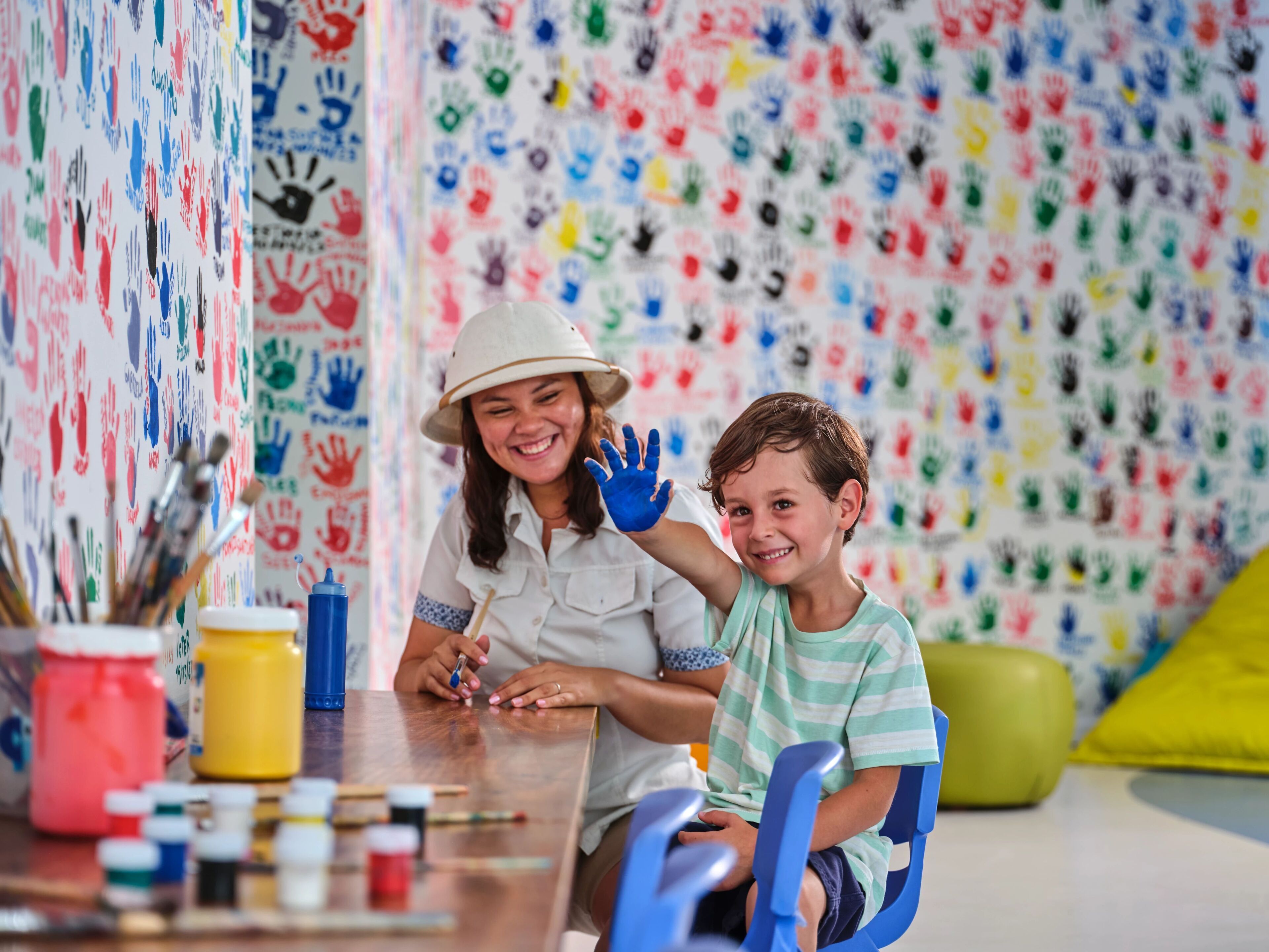 A room with a wall covered in colorful handprints, a table with art supplies, and a child and adult engaged in painting.