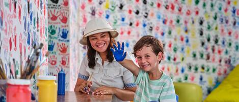 A room with a wall covered in colorful handprints, a table with art supplies, and a child and adult engaged in painting.