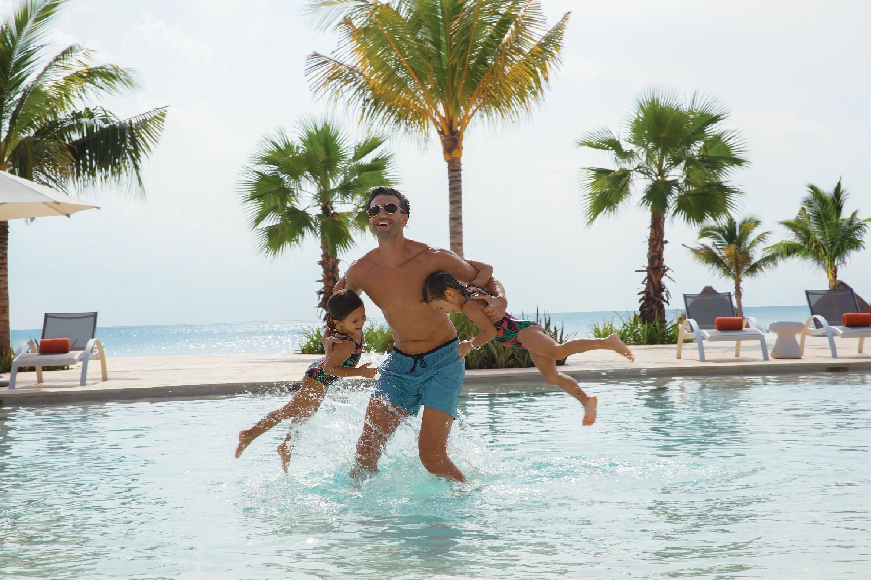 A man lifting a child out of a pool at a resort with palm trees and a beach view.