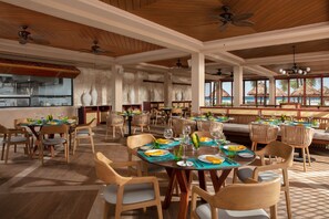 A spacious dining area with multiple tables and chairs, large windows with a view, and ceiling fans. - Dreams Aventuras Riviera Maya - All Inclusive (Puerto Aventuras)