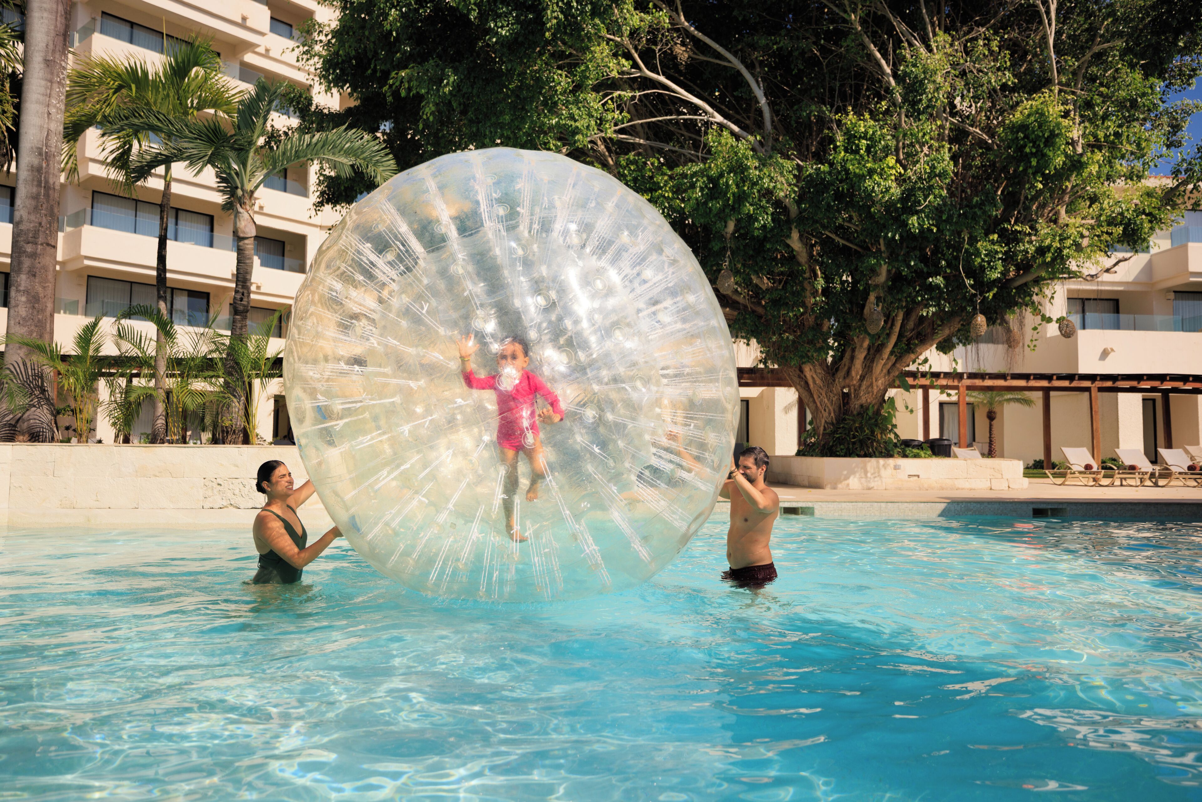 Una piscina con gente en el agua, una pelota inflable grande y un edificio con balcones y palmeras al fondo.