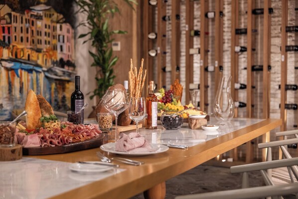 Une longue table à manger, dressée avec une variété de plats, des bouteilles de vin, et une fresque murale représentant un paysage en arrière-plan.