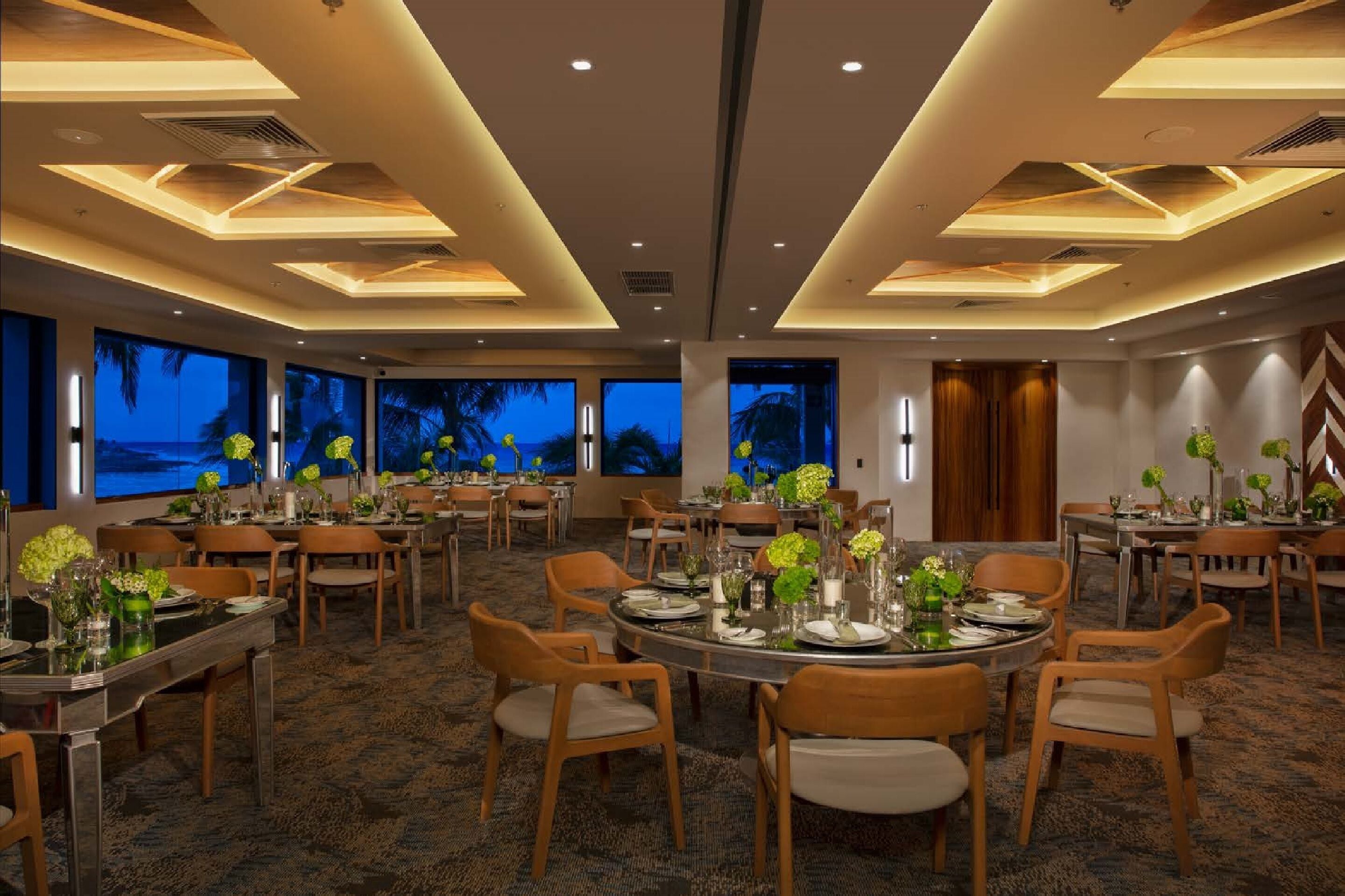 A spacious dining area with multiple tables and chairs, large windows with a view, and modern ceiling design.