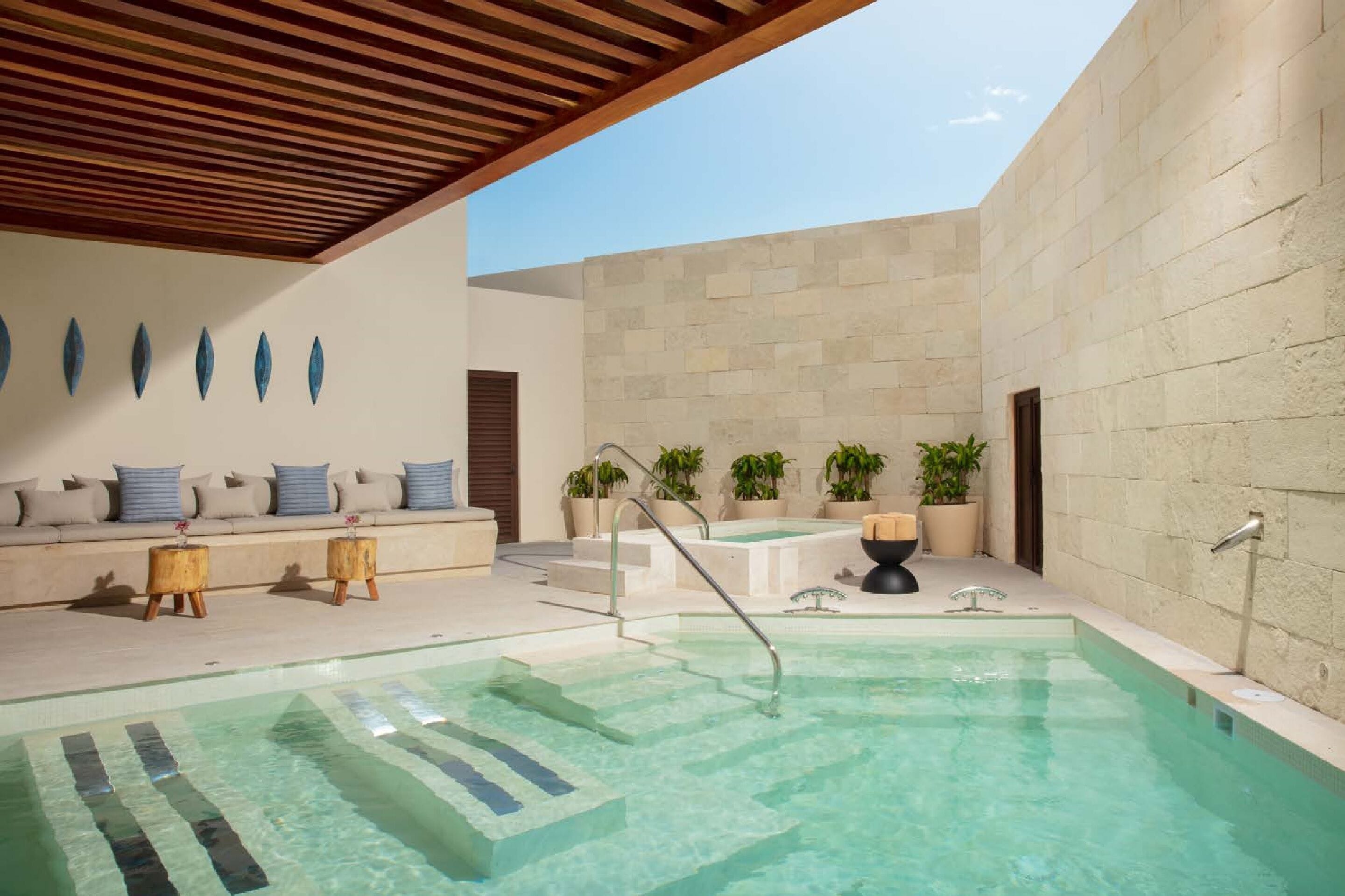 A pool area with a wooden ceiling, a seating area with cushions, and potted plants.