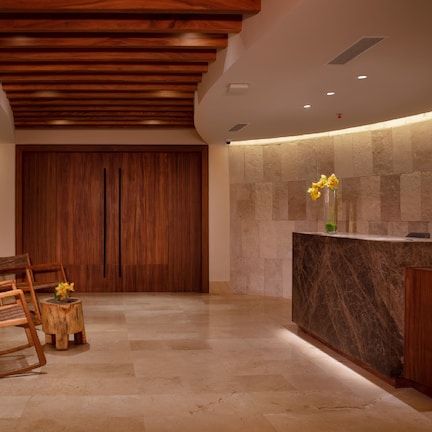 A reception area with a wooden reception desk, a stone wall, and a wooden door. There are two wooden chairs and a small round table with a vase of flowers.