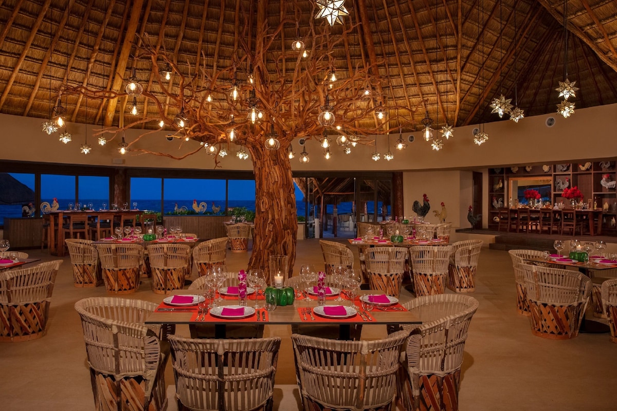 A restaurant with a large tree centerpiece, wicker chairs, and tables set for dining. - Dreams Aventuras Riviera Maya - All Inclusive (Puerto Aventuras)