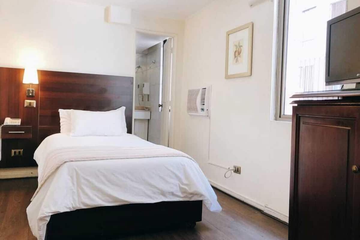 A hotel room with a bed, bedside table, lamp, air conditioning unit, and a television on a cabinet.
