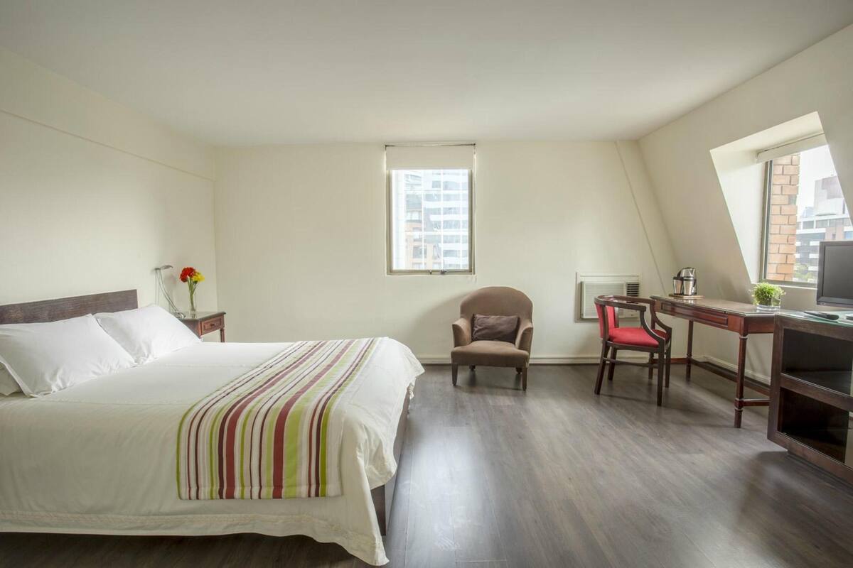 A bedroom with a bed, a desk, a chair, a window, and a TV.