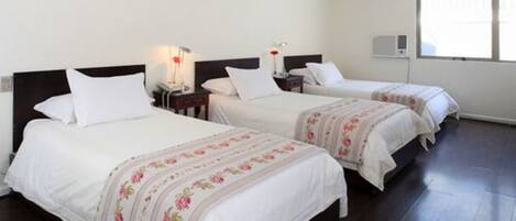 Quarto de hotel com duas camas, cada uma com roupa de cama branca e estampas florais. Apresenta uma mesa de cabeceira com uma luminária e uma mesinha com um vaso de flores vermelho. Uma janela oferece iluminação natural.