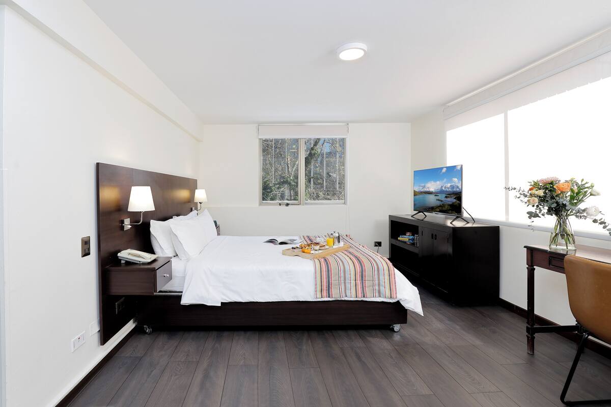 A modern hotel room with a large bed, bedside lamps, a desk with a computer, a TV, and a window with a view of trees.