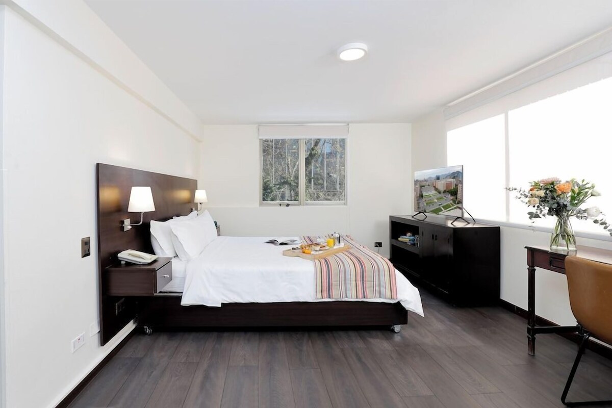 A modern hotel room with a bed, bedside lamps, a desk, and a window with a view of trees.