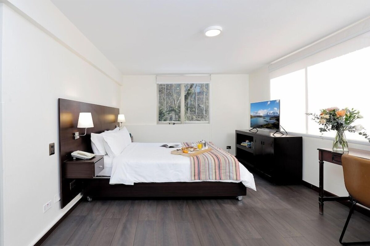A modern hotel room with a bed, bedside lamps, a TV, a desk with a chair, and a window with a view of trees.