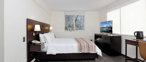Habitación de hotel con cama, escritorio, silla, televisor y ventana con vista a árboles.