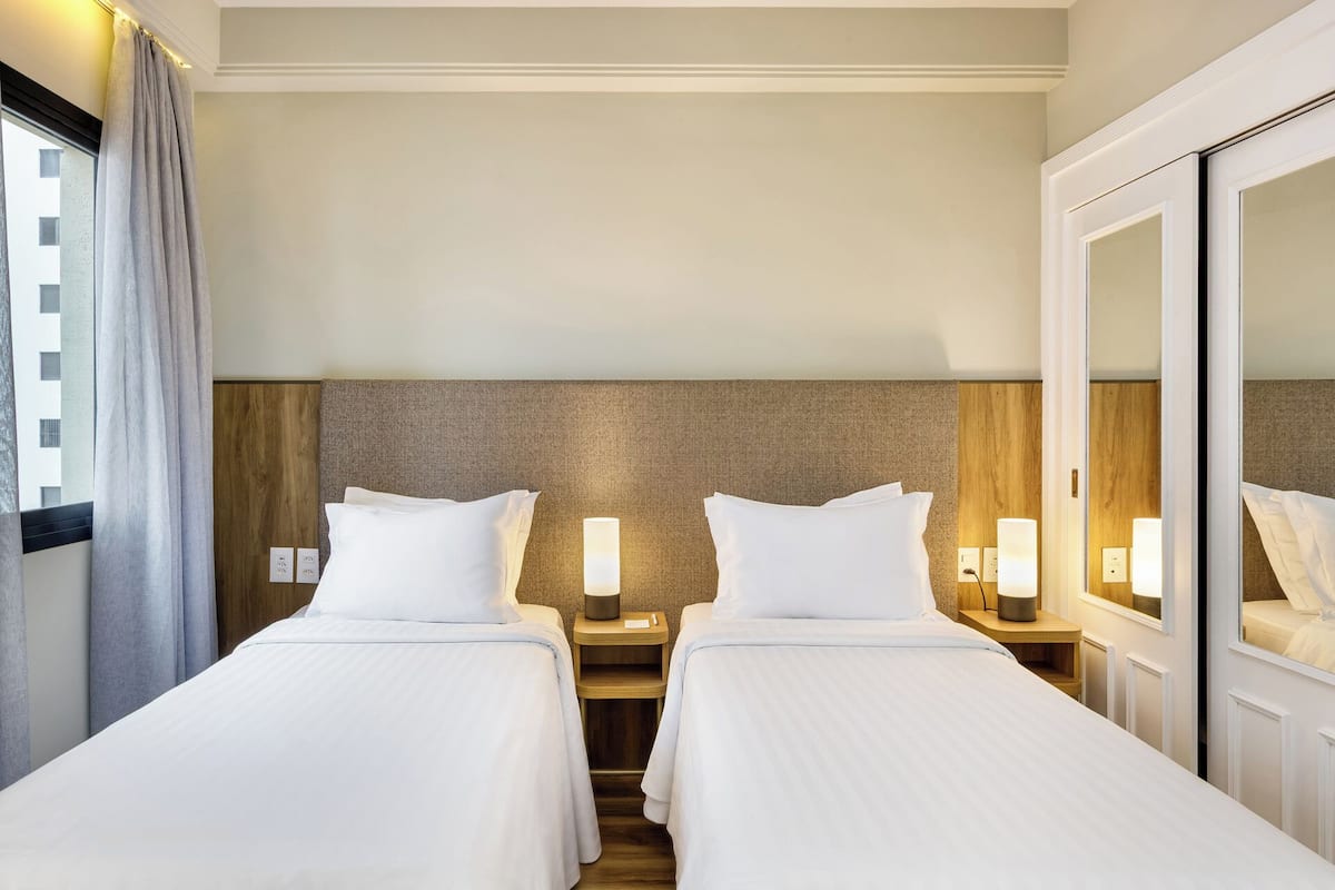 Une chambre d’hôtel avec deux lits, des têtes de lit en bois, des lampes de chevet, un miroir et une vue sur un bâtiment à l’extérieur.