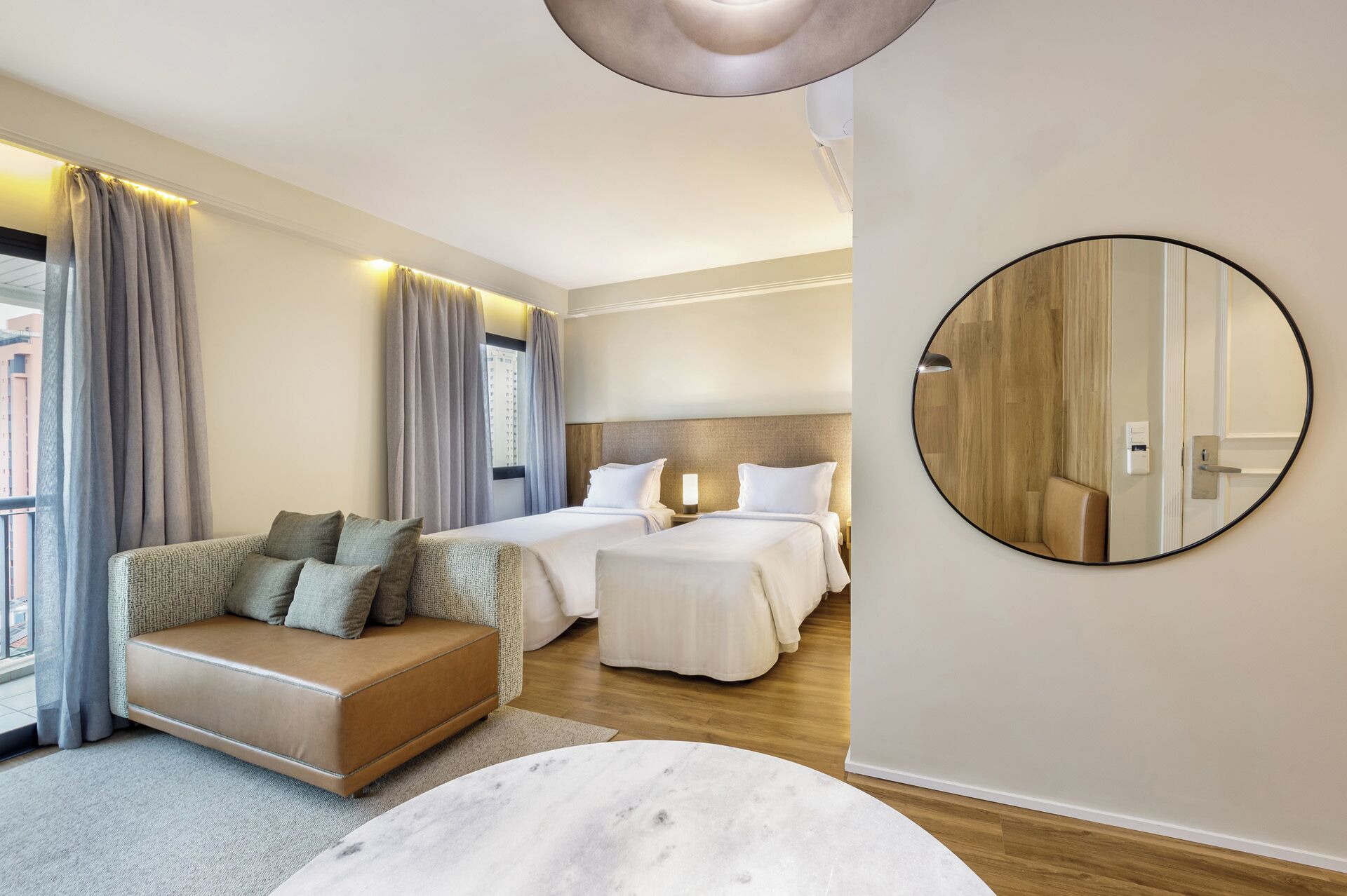 A hotel room with a sofa, two beds, a round mirror, and a view of the city.
