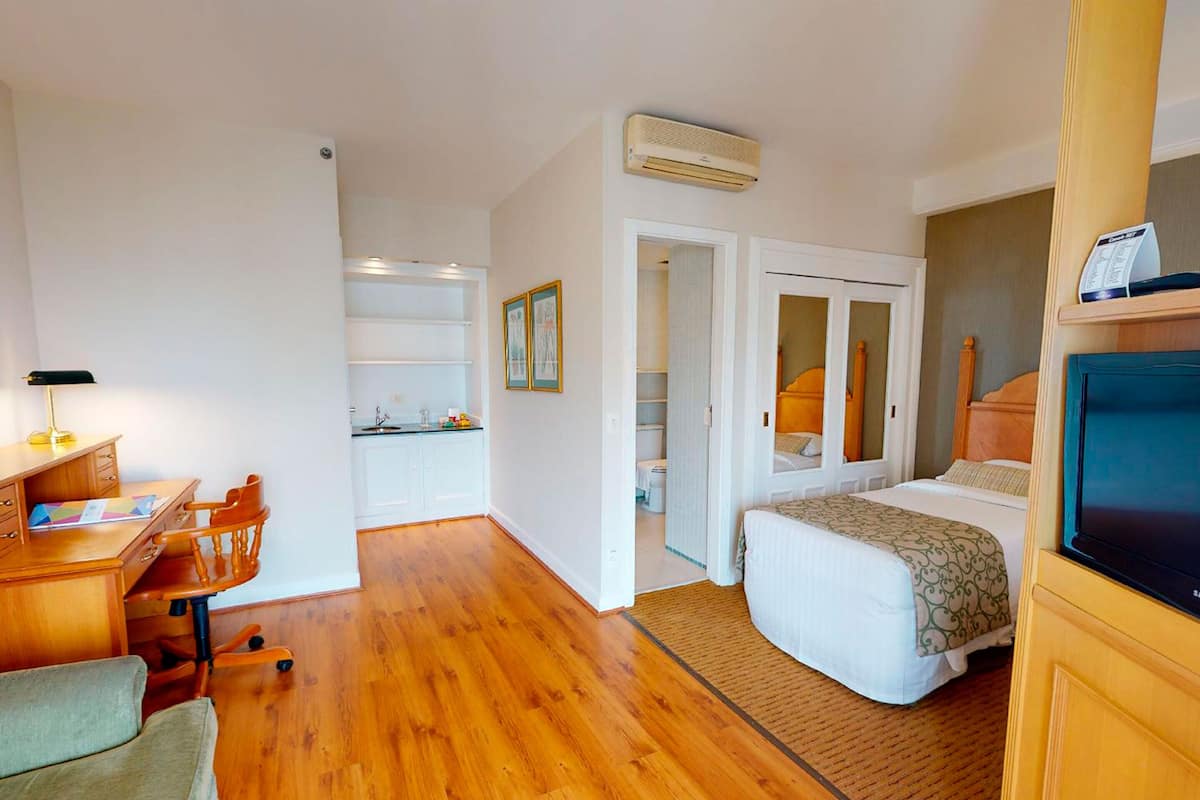 Une chambre d’hôtel avec un plancher en bois, un bureau avec une chaise, un lit recouvert d’une couette blanche, une télévision et une armoire.