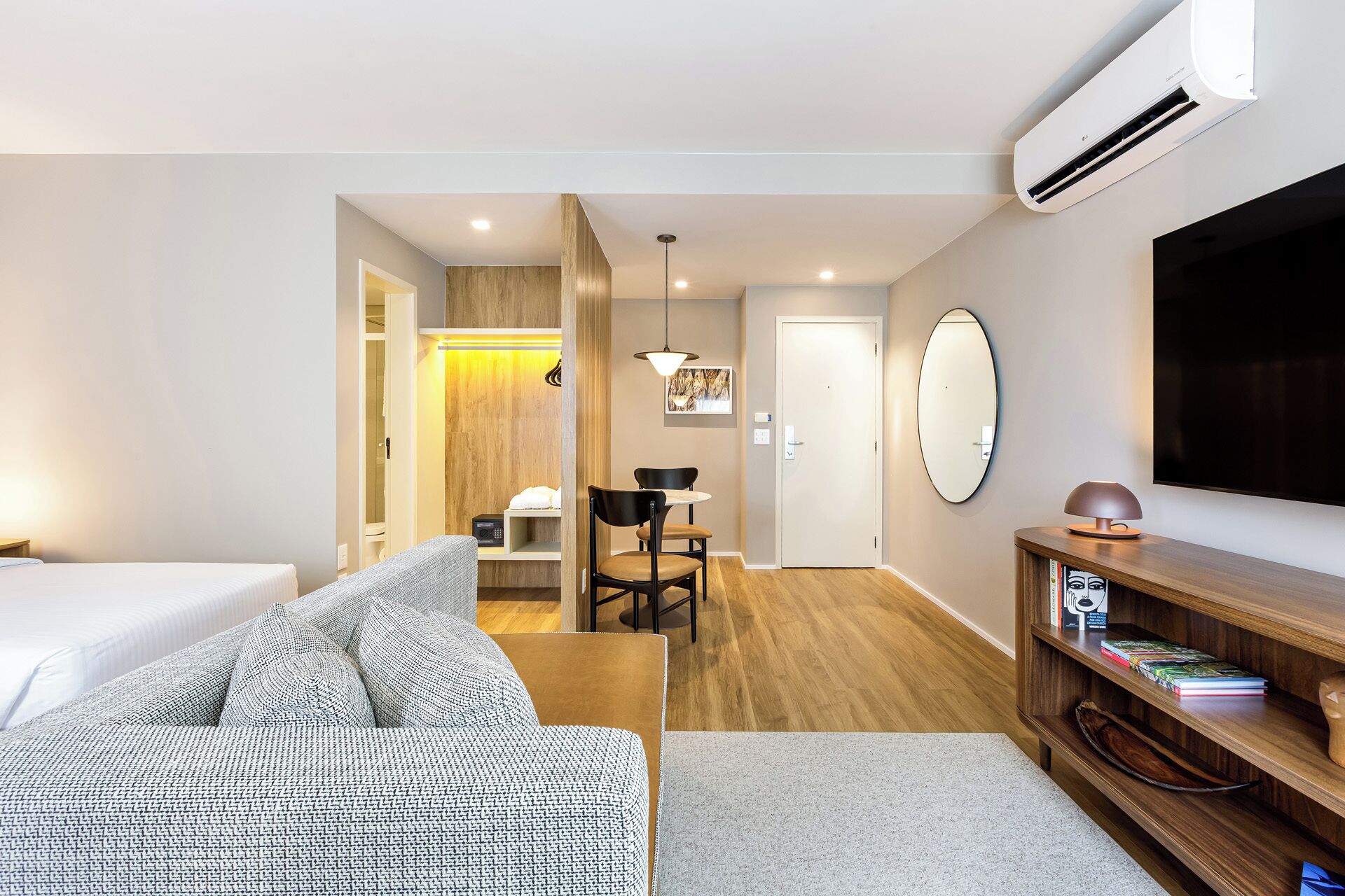 A hotel room with a bed, a sofa, a desk with a chair, a TV, a bookshelf, and a mirror.