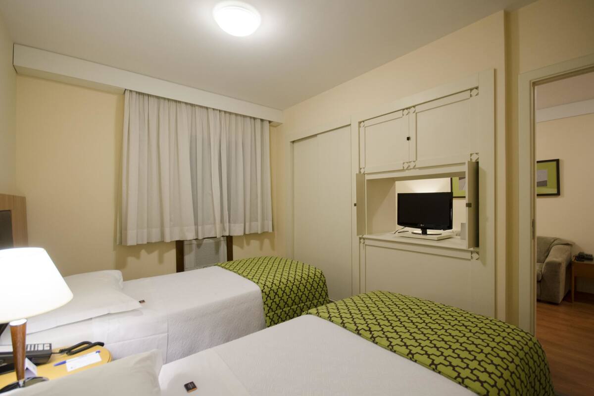 Habitación de hotel con dos camas individuales, un televisor montado en un mueble, un escritorio con lámpara y una ventana con cortinas.