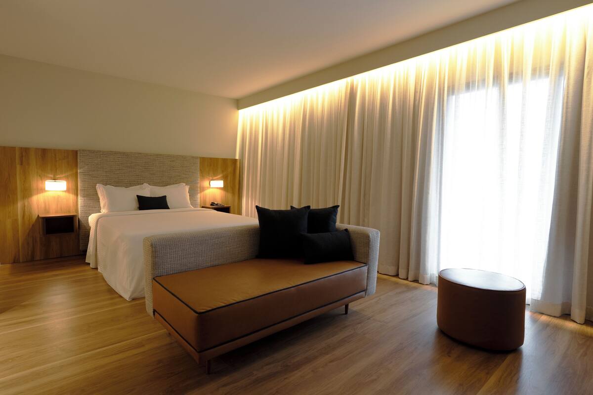 Una habitación de hotel moderna con una cama grande, un sofá, un reposapiés redondo y una mesita de noche con lámpara.
