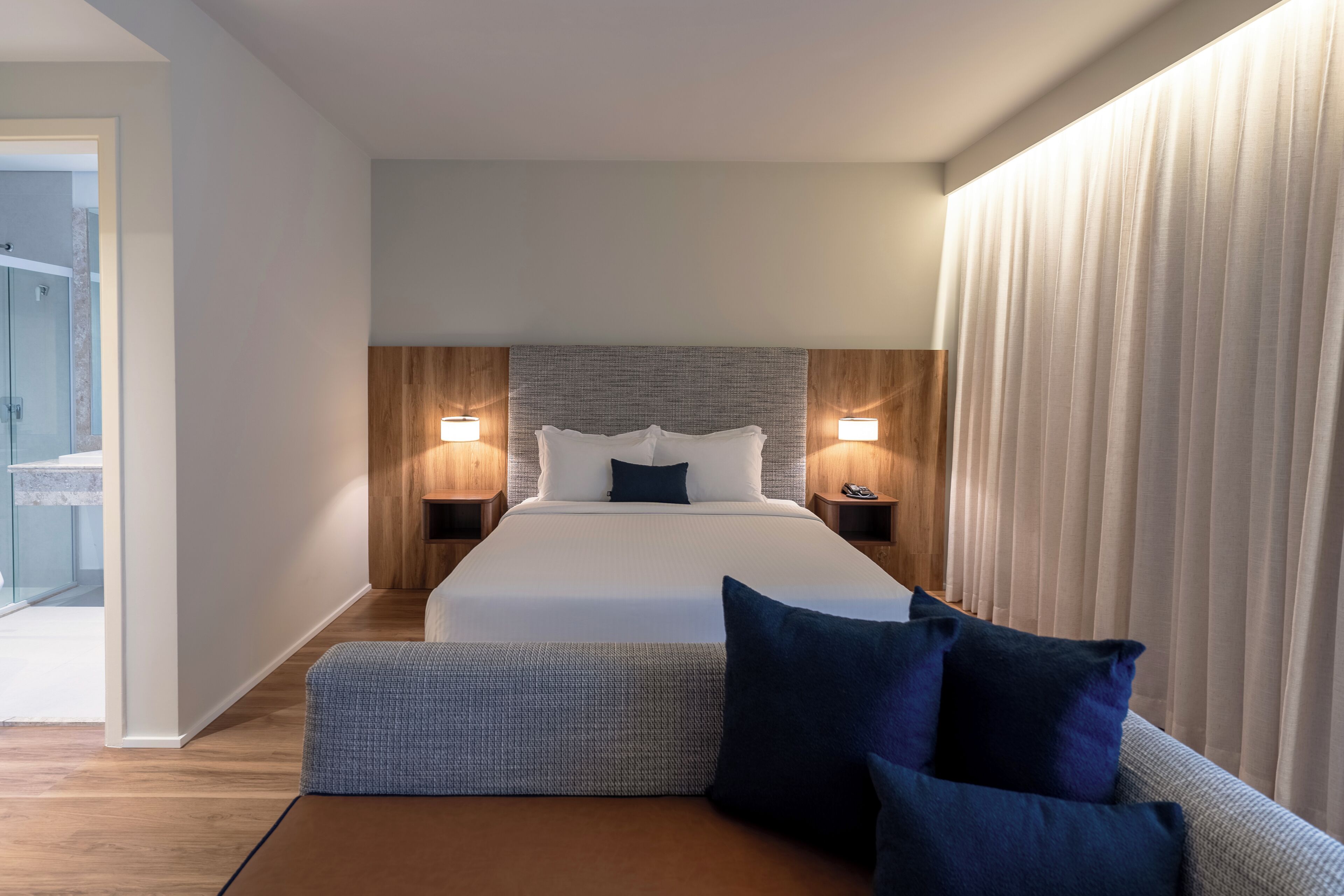 A modern hotel room with a large bed, bedside lamps, a bench, and a view of the hallway.