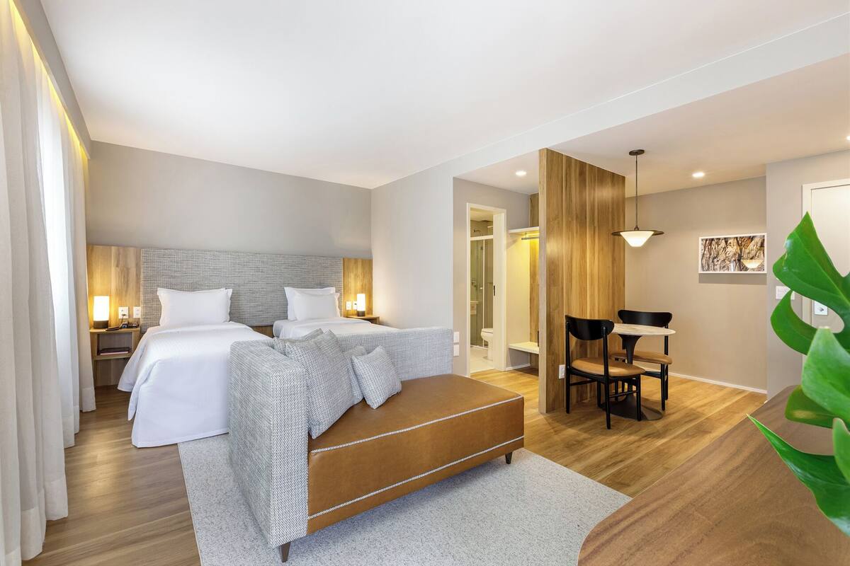 Habitación de hotel con dos camas, una zona de estar con mesa y sillas, y un área de ducha.