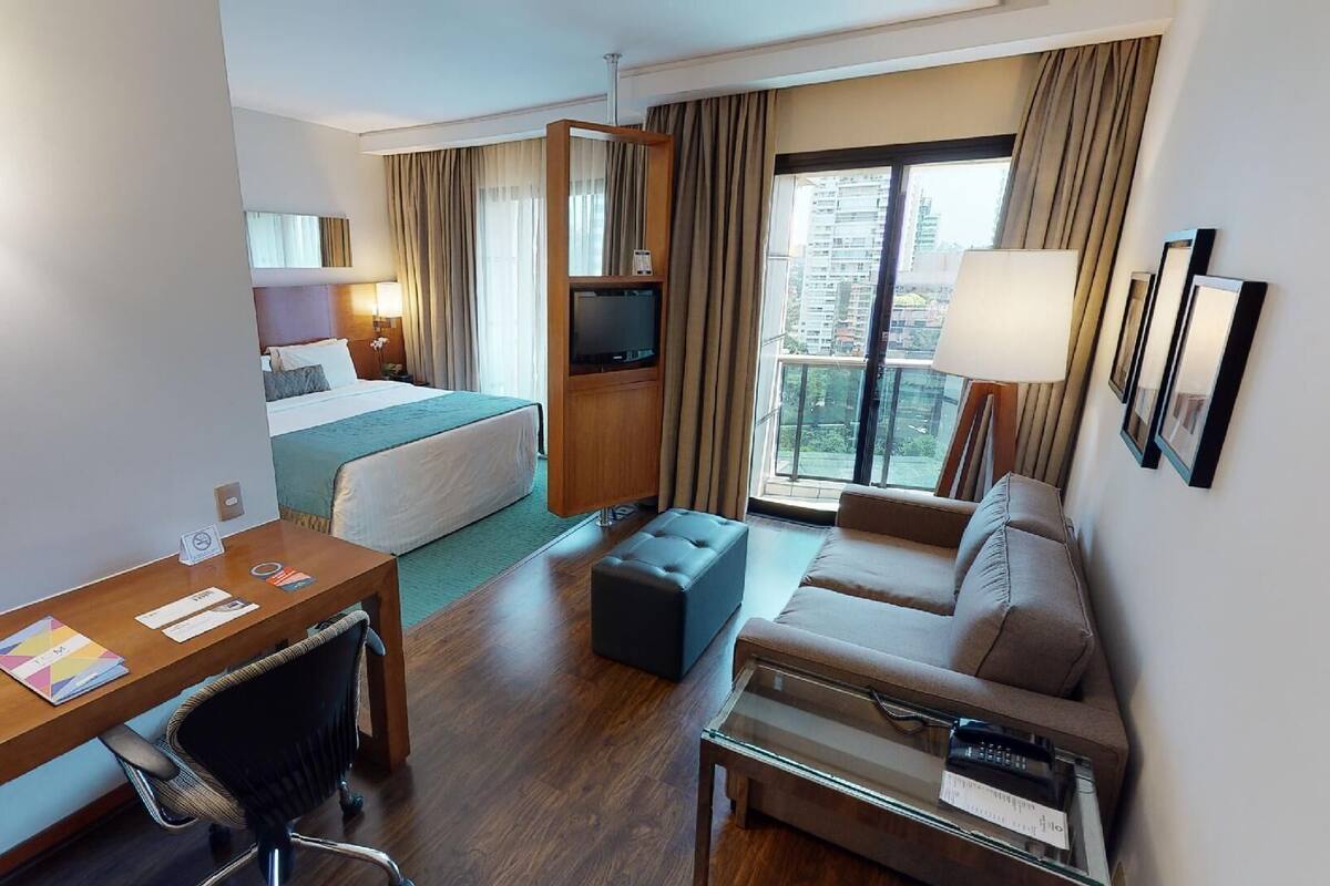 A hotel room with a bed, a desk, a sofa, a TV, and a large window with a city view.