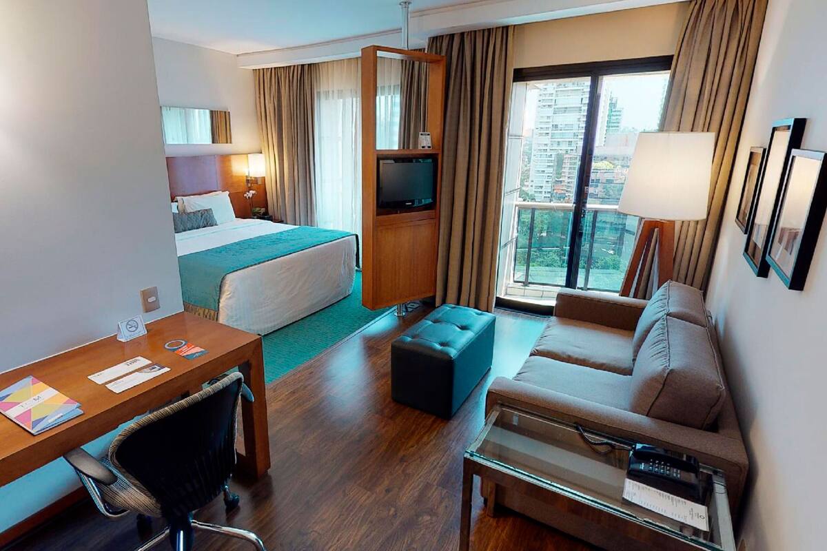 A hotel room with a bed, a sofa, a desk, a TV, and a balcony with a city view.
