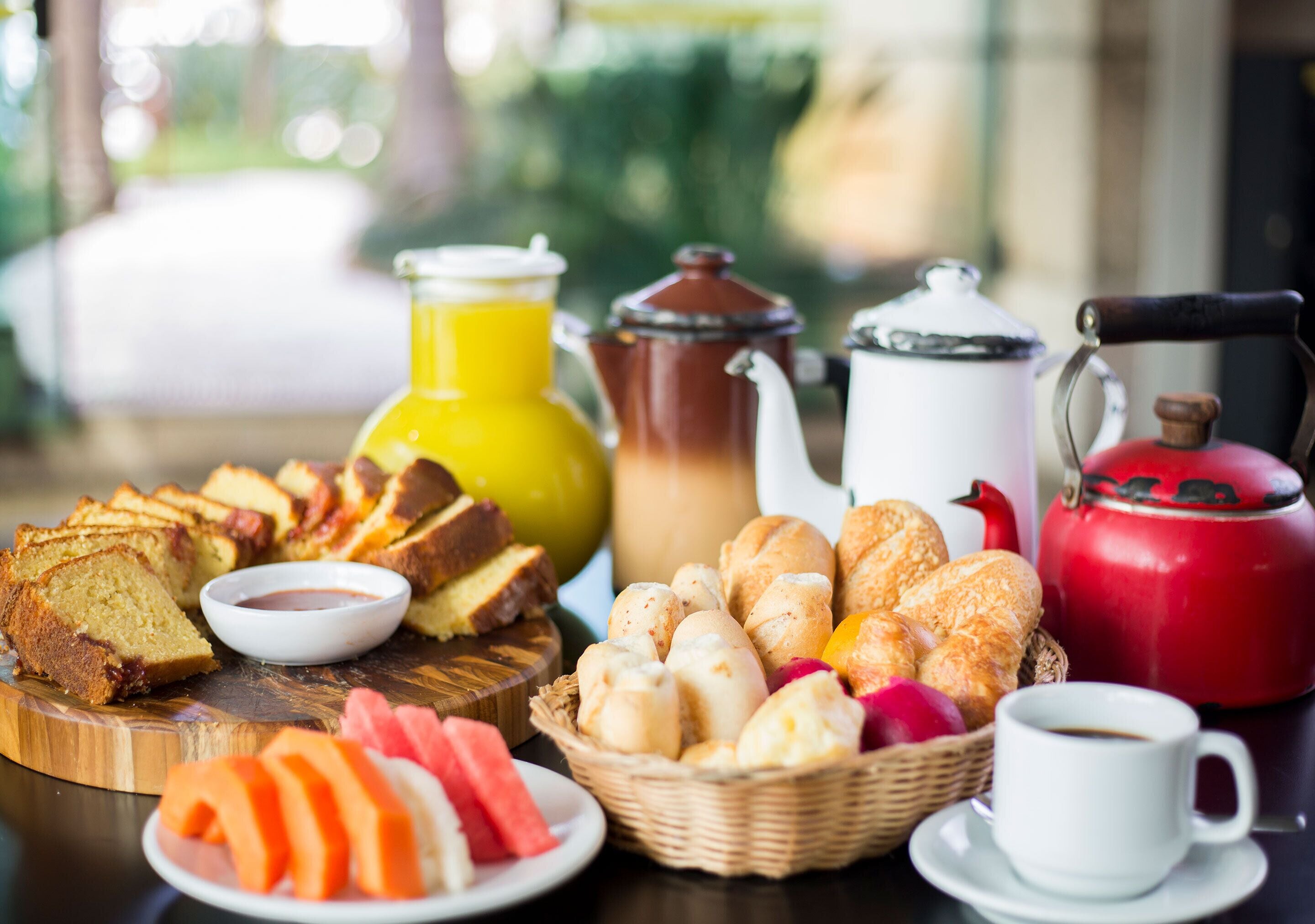 A breakfast table with various pastries, bread, a jug of orange juice, a teapot, a cup of coffee, and a red kettle.