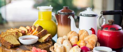 A breakfast table with various pastries, bread, a jug of orange juice, a teapot, a cup of coffee, and a red kettle.