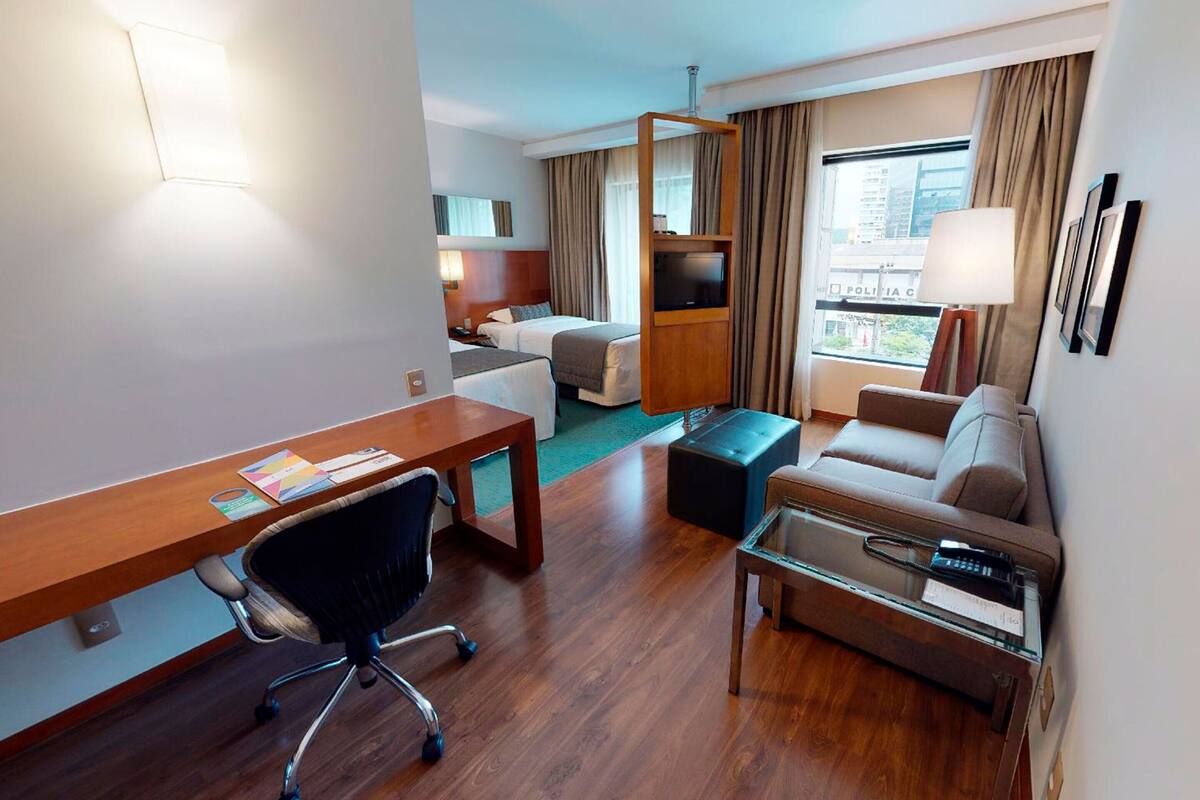 A hotel room with a wooden desk, a black office chair, a sofa, a glass coffee table, and a TV on a wooden stand.