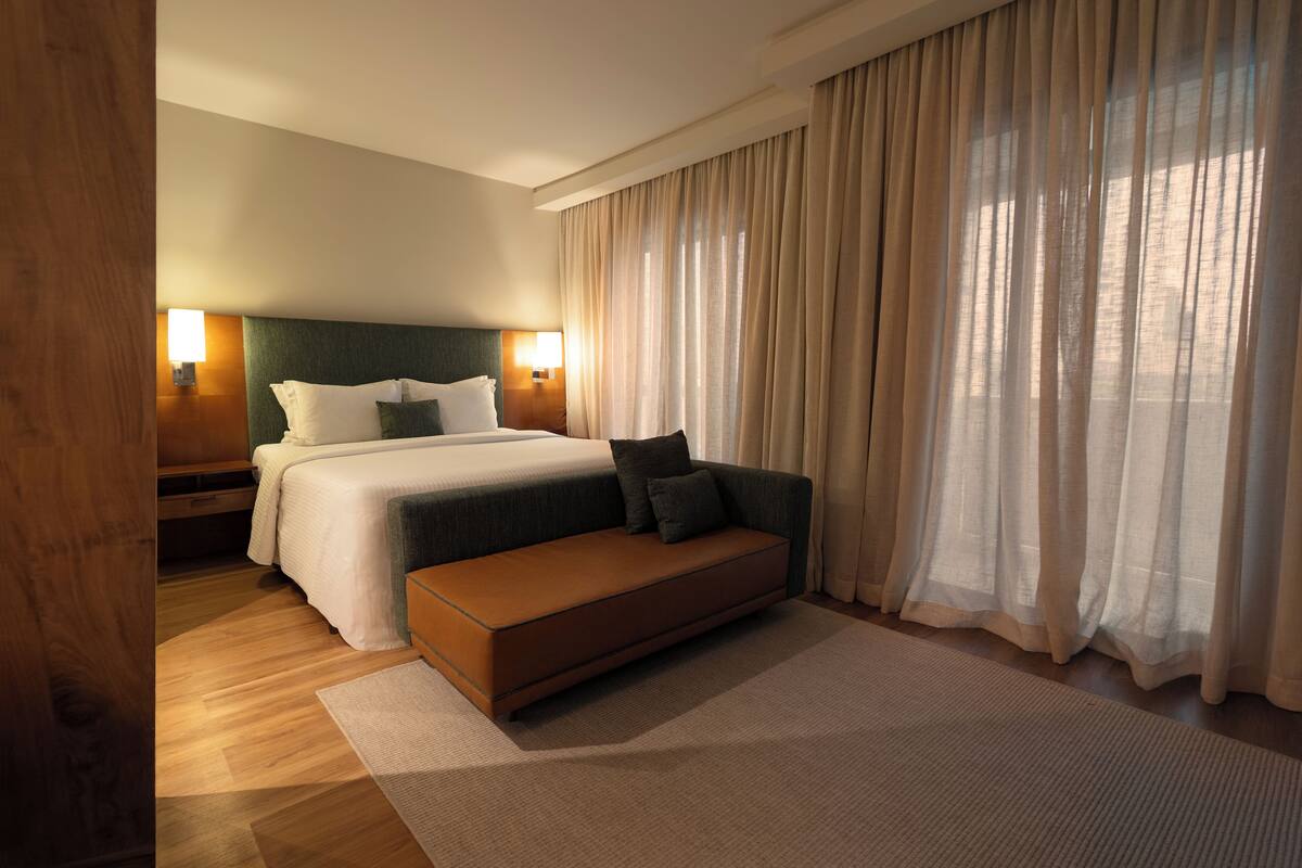 A modern hotel room with a bed, a sofa, a bench, and a large window with curtains.