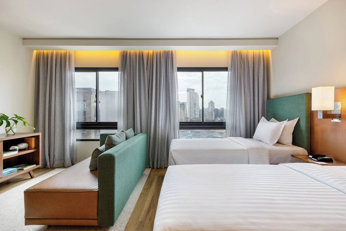A hotel room with two beds, a sofa, a small table, and a view of the cityscape.