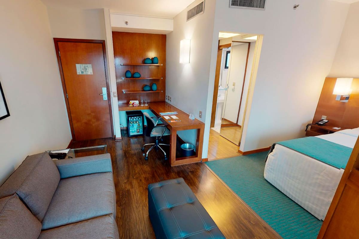 A hotel room with a sofa, a small table, a desk with a chair, a bed, and a wooden shelf with decorative items.