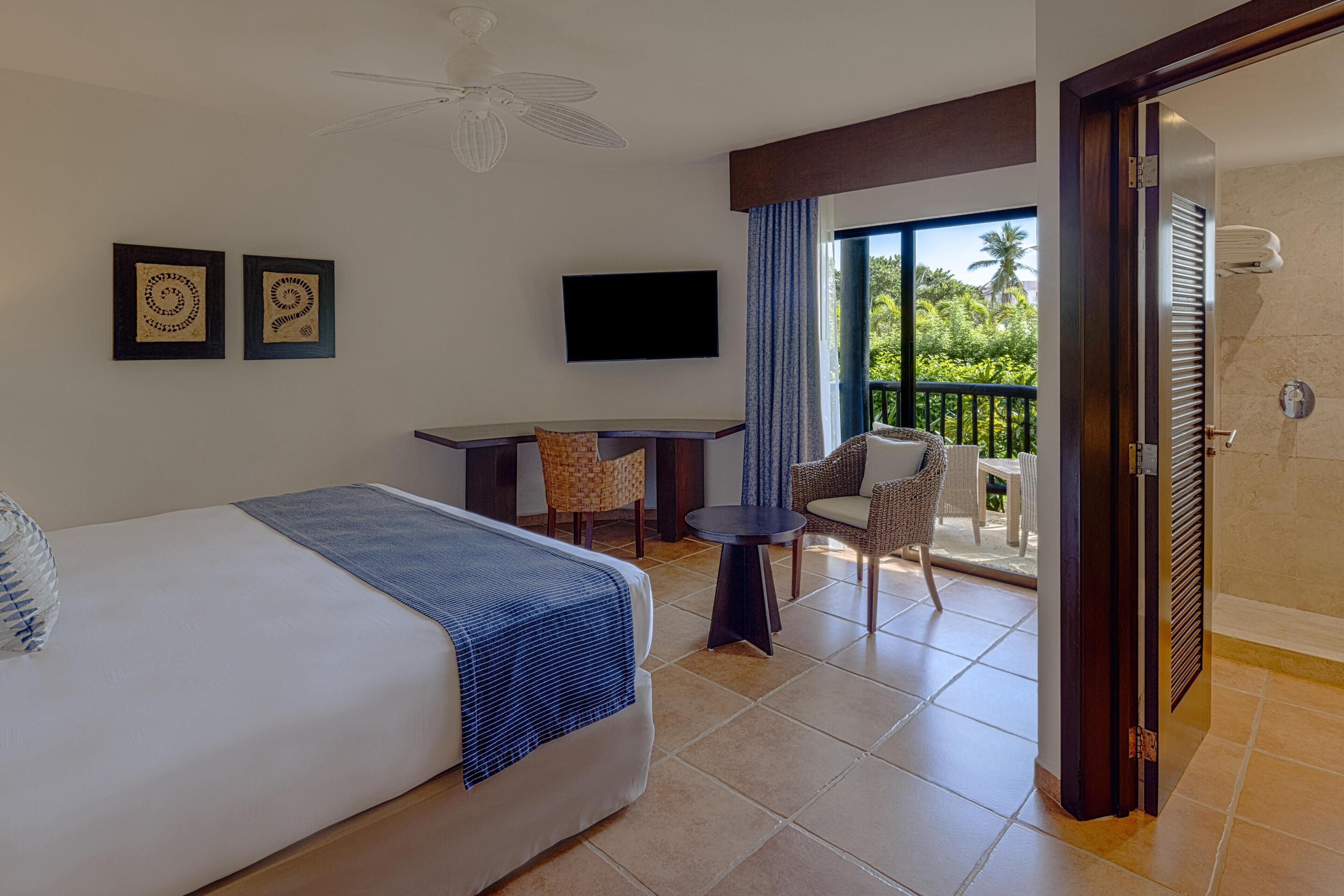 A hotel room with a bed, a desk, a TV, a balcony with chairs, and a ceiling fan.