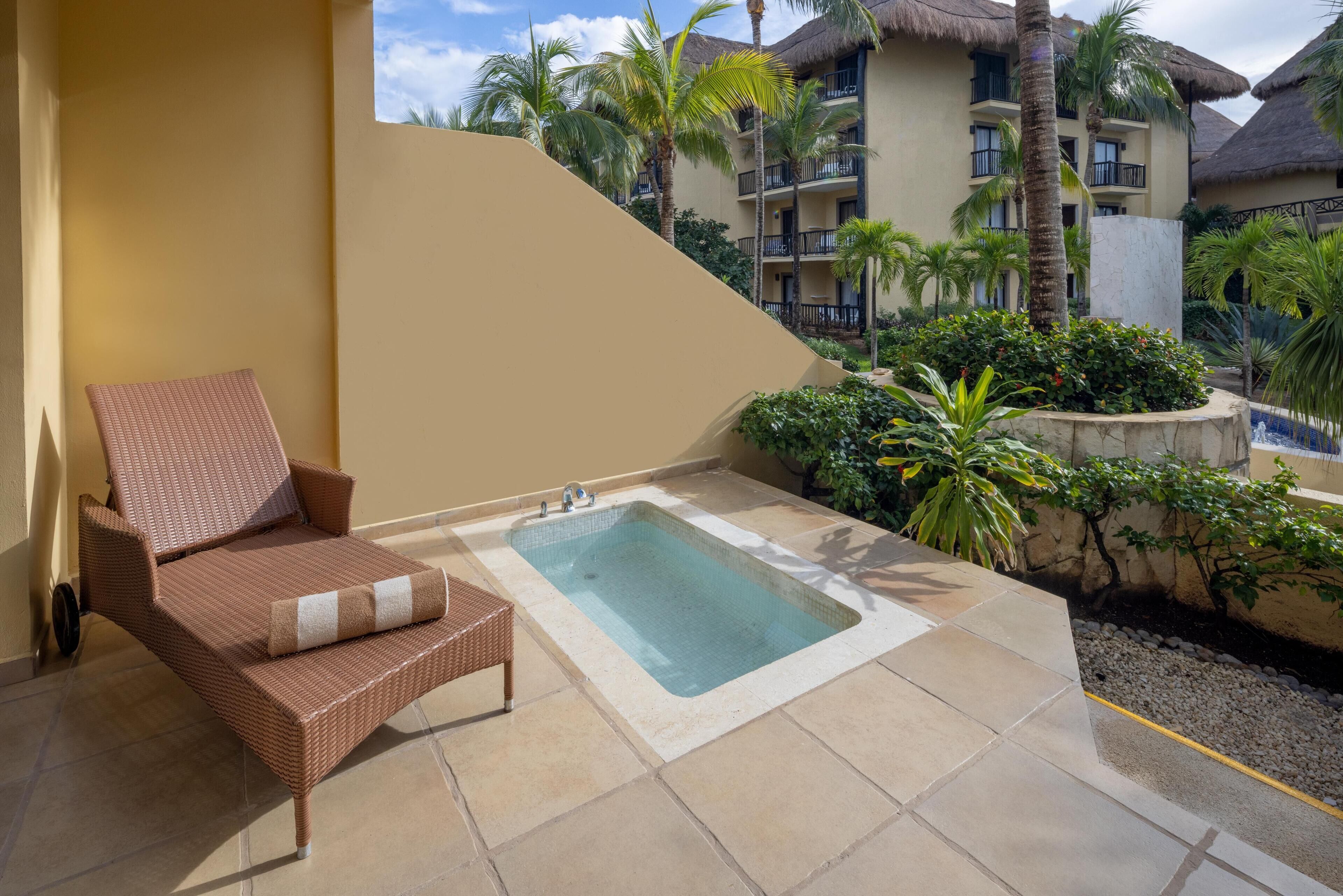 Eine Lounge-Ecke mit Korbstuhl, kleinem Pool und Blick auf ein Resortgebäude mit Balkonen und Palmen.