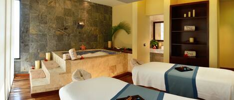 Habitación de hotel con pared de piedra, una bañera, dos camas, un estante con velas y una planta pequeña.