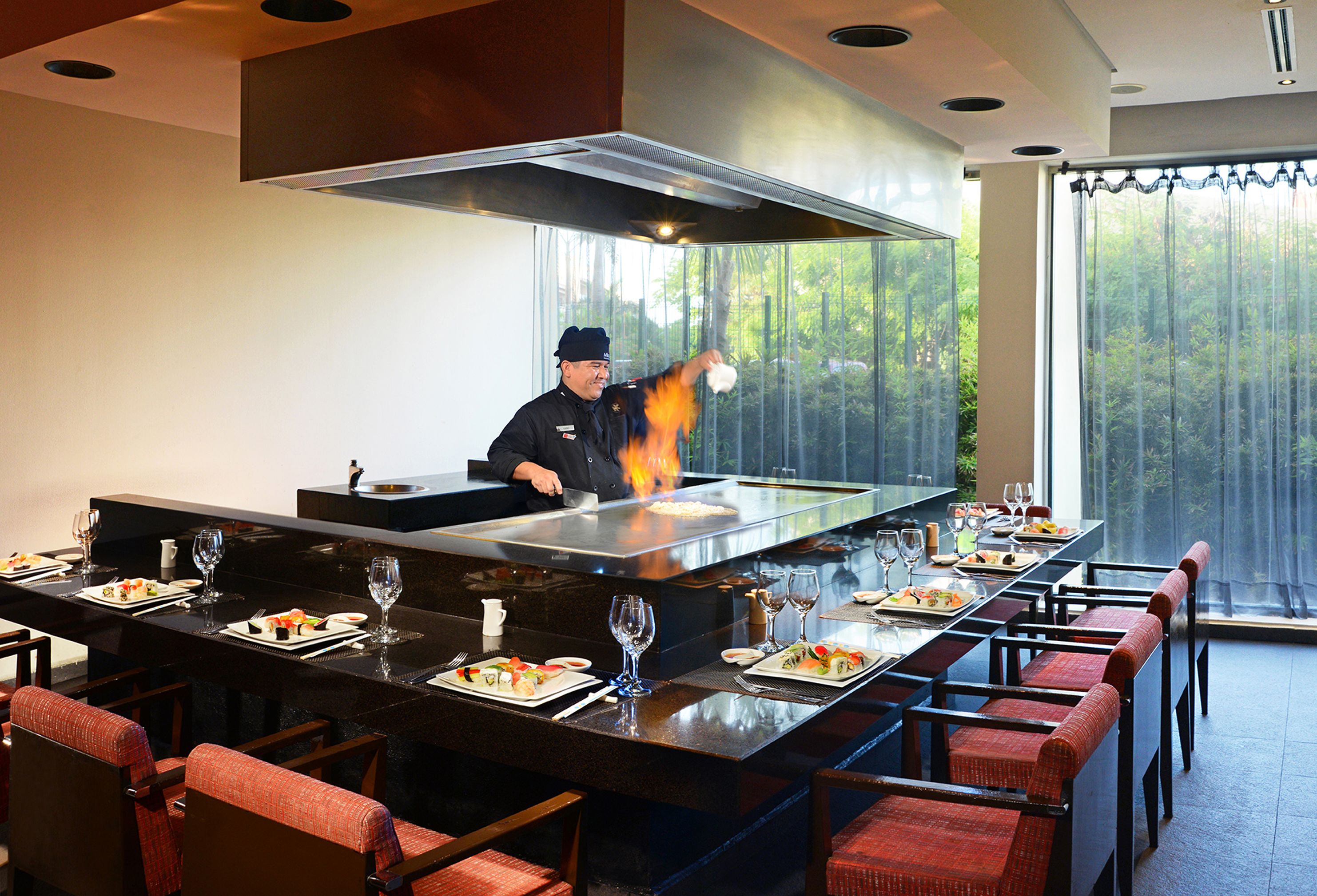 A restaurant with a chef cooking on a large stove, a long dining table with chairs, and a view of trees through the windows.