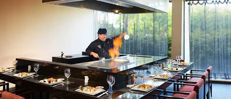 A restaurant with a chef cooking on a large stove, a long dining table with chairs, and a view of trees through the windows.