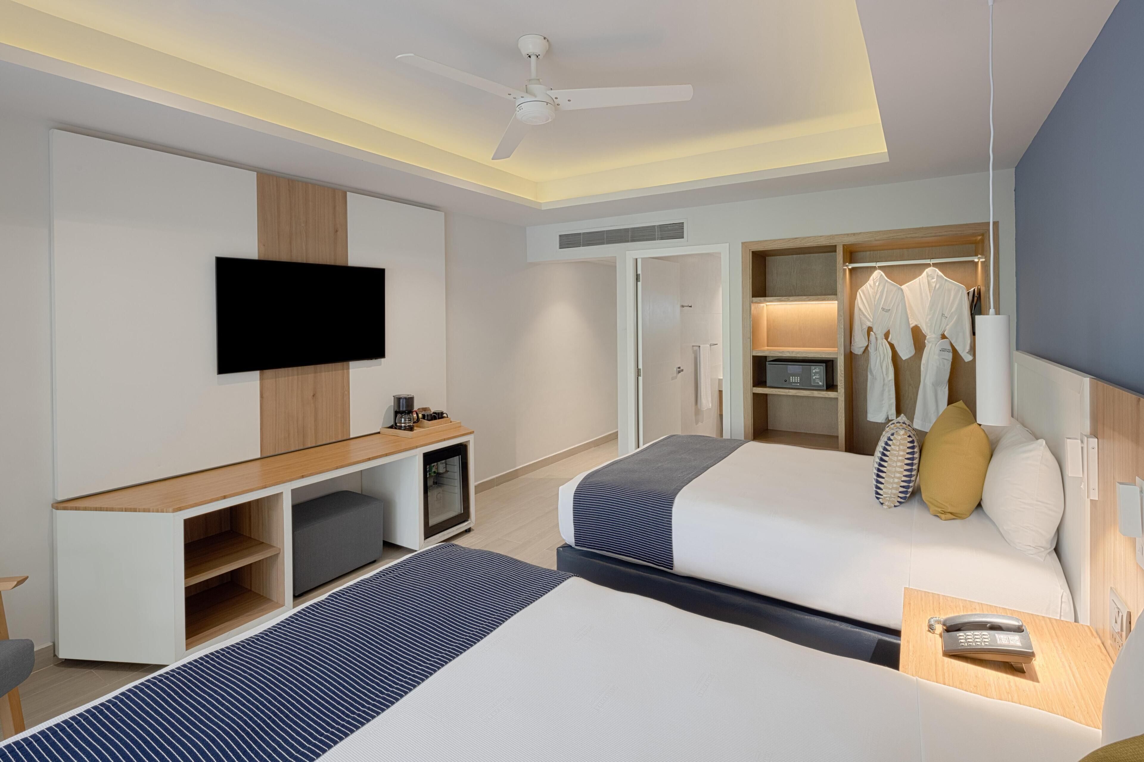 Una habitación de hotel moderna con dos camas, un televisor de pantalla plana, ventilador de techo, un minibar y vestidor.