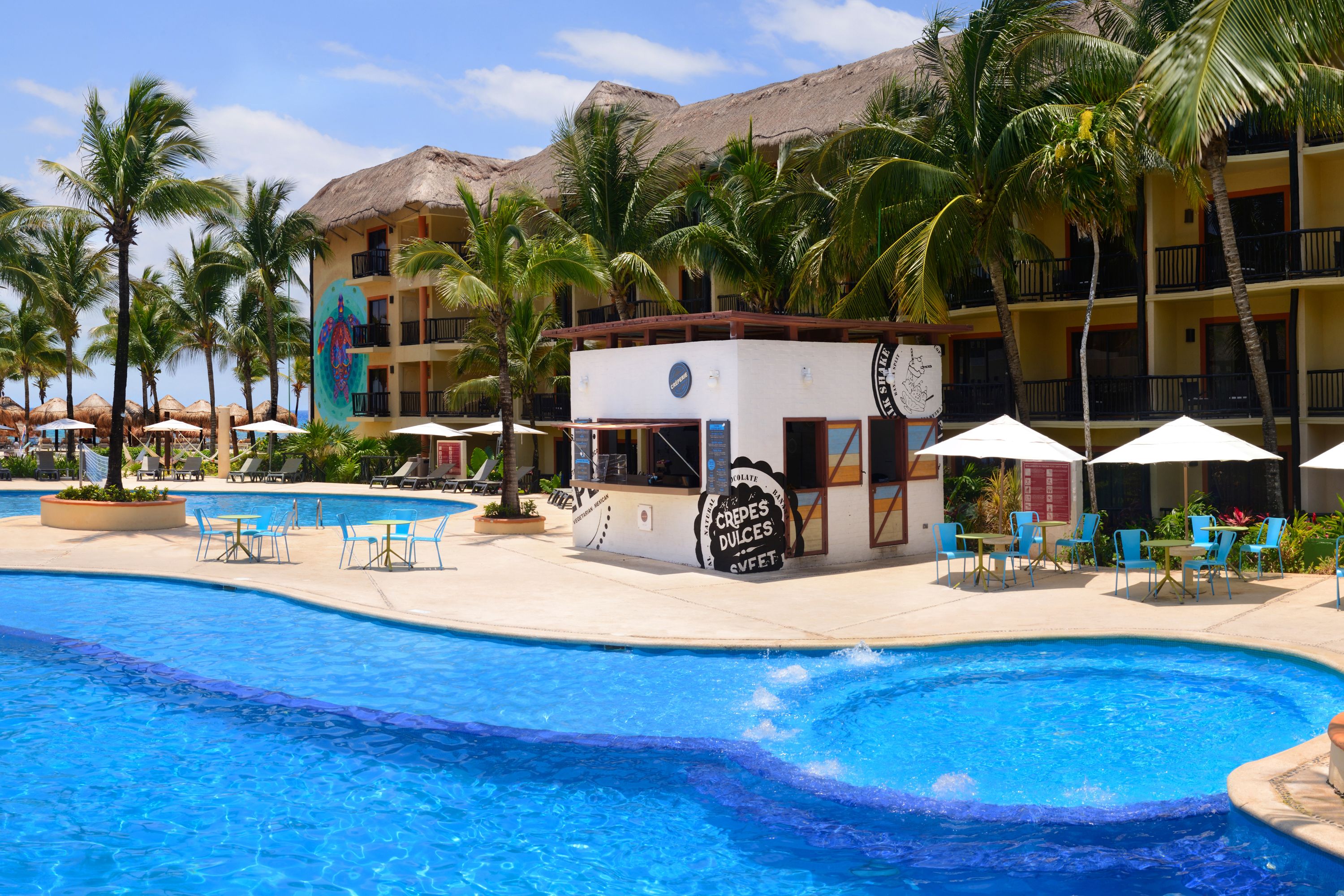 A resort pool area with a small building labeled 'CREDES DULCES SWEET', surrounded by palm trees and lounge chairs.