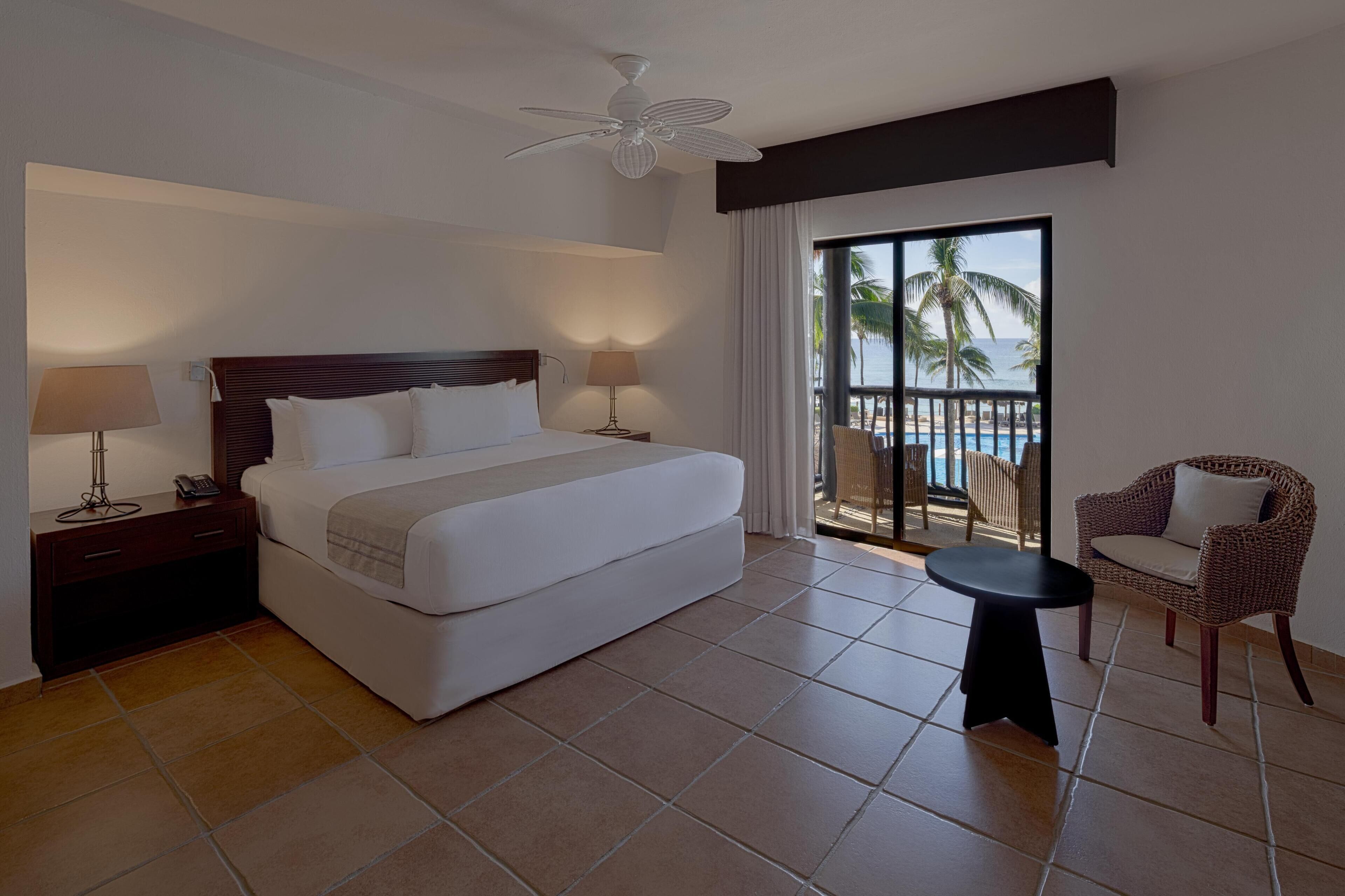 A hotel room with a bed, a nightstand, a lamp, a chair, a small table, a ceiling fan, and a balcony with a view of the beach and palm trees.