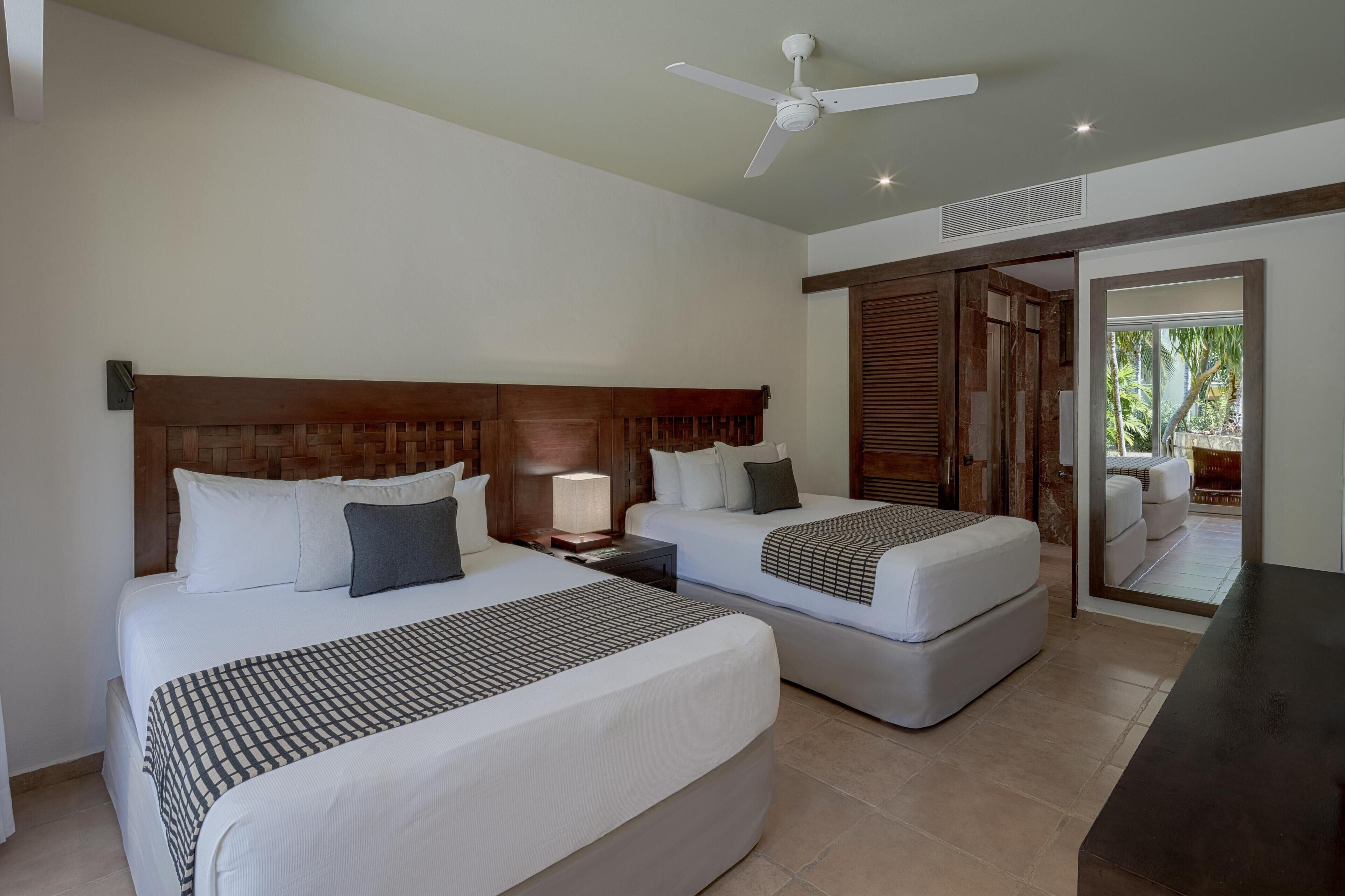 A hotel room with two beds, a ceiling fan, a nightstand with a lamp, and a view of the outside.