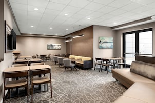 A hotel lobby with multiple seating areas, including tables and chairs, a sofa, and a large window providing natural light.