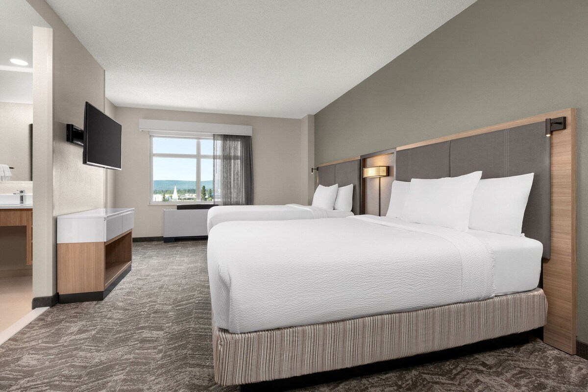 A hotel room with two beds, a TV, a desk, and a window with a view.