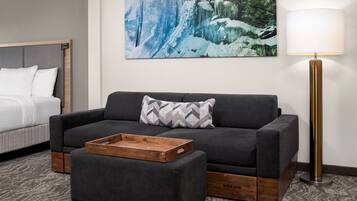 Une chambre d’hôtel moderne avec un canapé gris, une table d’appoint en bois, un lit blanc et un grand tableau mural représentant une cascade.